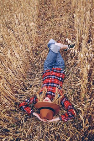 Blonde woman in plaid shirt with fedora hat lying on brown grass field