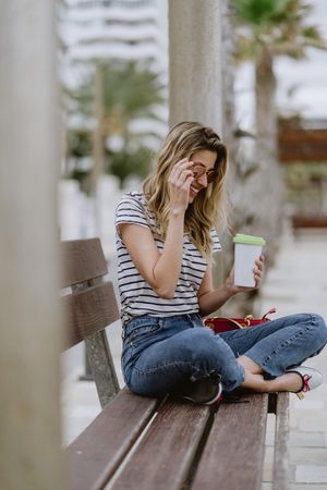 Happy woman with disposable cup of coffee sitting on street bench