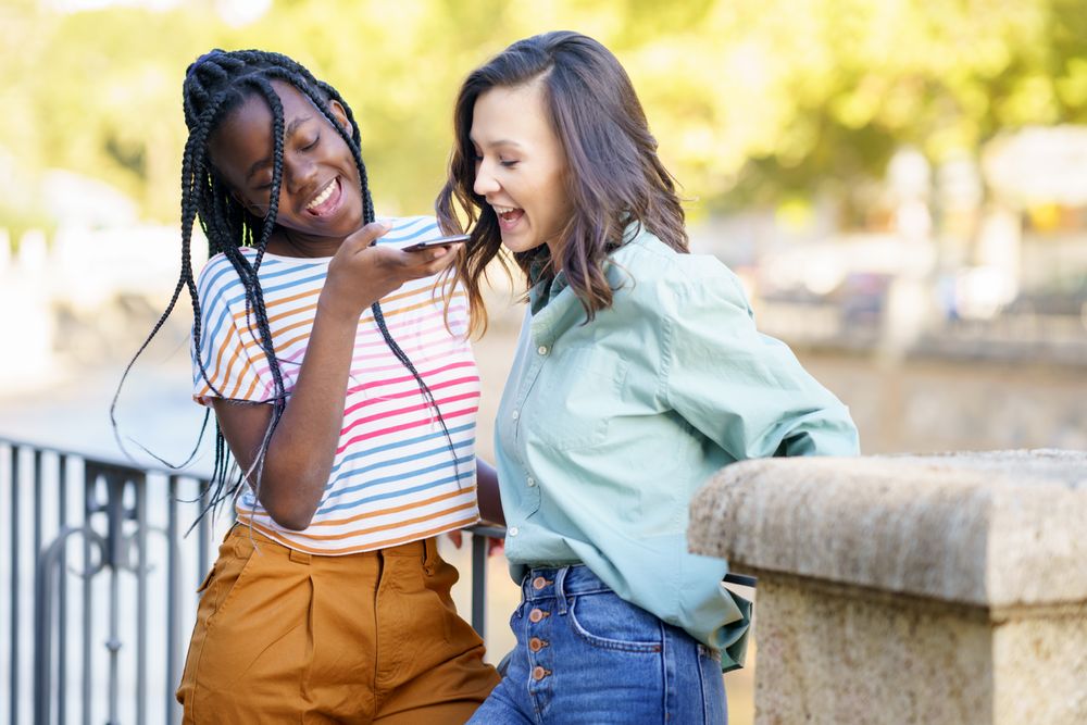 Female friends smiling while using speaker phone in casual clothing outside