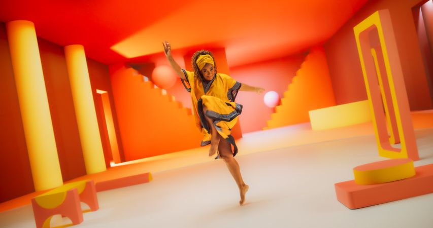 Black Woman in African Outfit Dancing Energetically in Geometric Abstract Orange Environment. Creative Female Performing Dance Choreography in a Studio While Looking at the Camera.