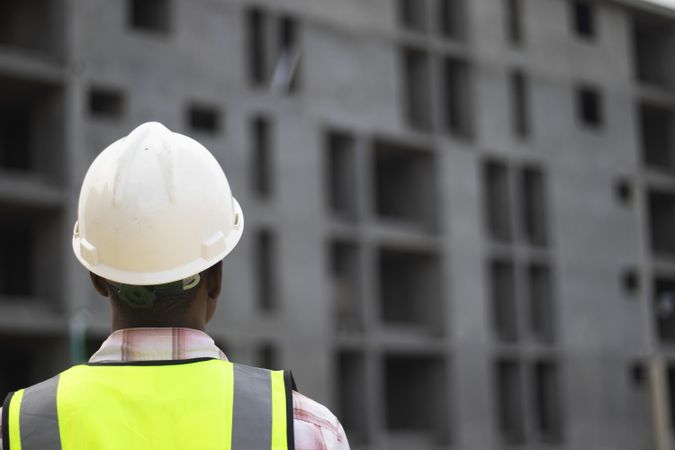 Back view of man wearing vest and bump cap standing beside a building under construction