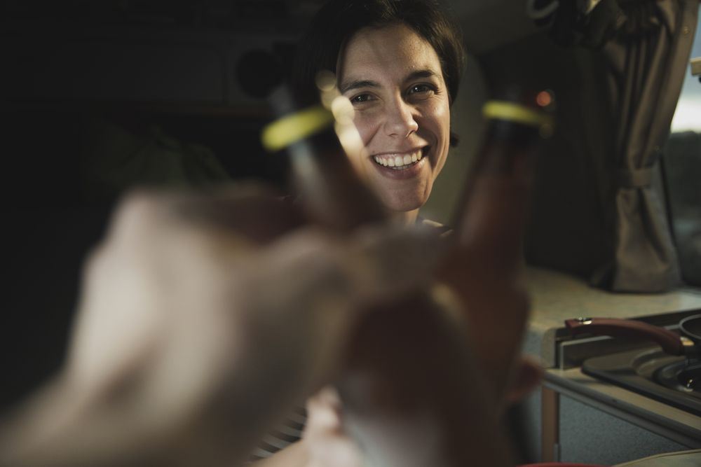 Smiling woman clanging bottles with person near cooker in camper van
