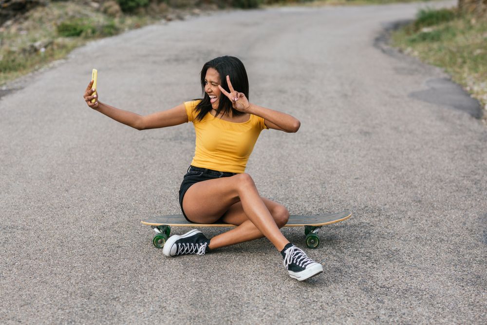 Delightful Latina woman on longboard taking selfie on smartphone
