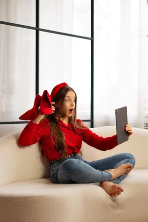 Surprised teen girl holding red shoes making video call.