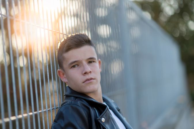 Teenage male wearing a leather jacket leaning on fence outside looking at camera