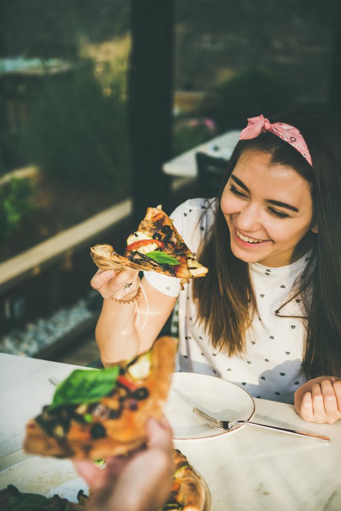 Couple eating pizza al fresco with wine, woman smiling holding pizza