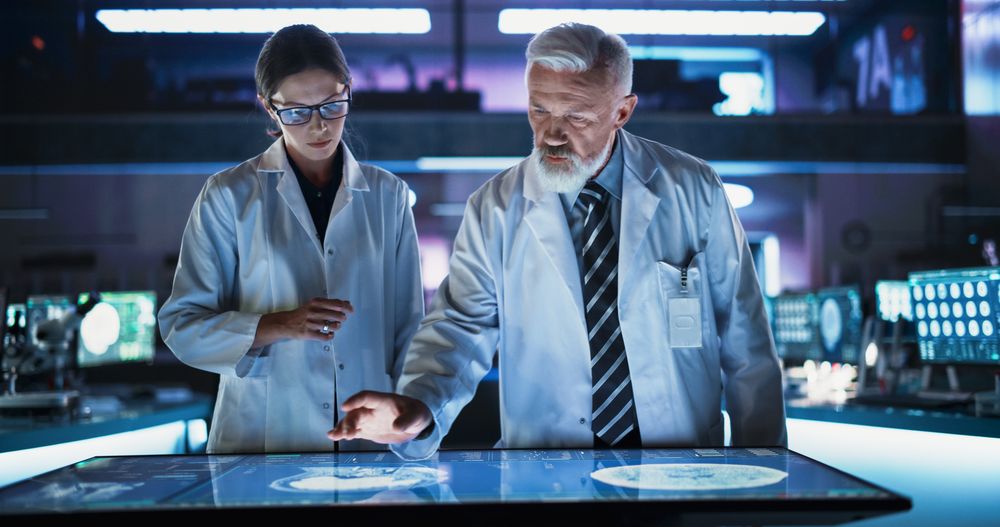 White Female Neuroscientist And Male Surgeon Talking, Using Interactive Touch Screen Table With CT Scan Of Human Brain With Tumor, Finding Treatment
