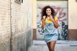 Arab woman with headphones and curly hairstyle walking outdoors in lane with graffiti