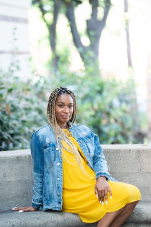 Portrait of woman in yellow dress and jean jacket sitting outside on bench smiling