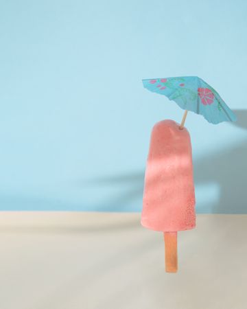 Pink ice pop on blue background with sand with blue parasol