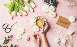 Coffee, tulips artfully arranged on pink background, with “happy” card, and hands on coffee