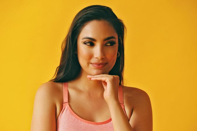 Headshot of calm Hispanic woman looking to her side with hand up to her chin in a yellow background