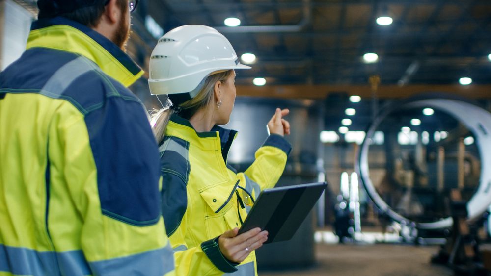 Male and Female Industrial Engineers in Hard Hats and Safety Jackets Discuss New Project while Using Tablet Computer.