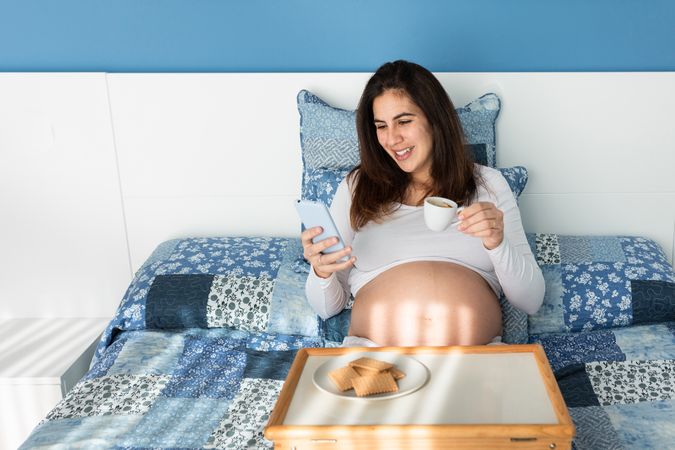 Happy pregnant woman having breakfast and browsing smartphone