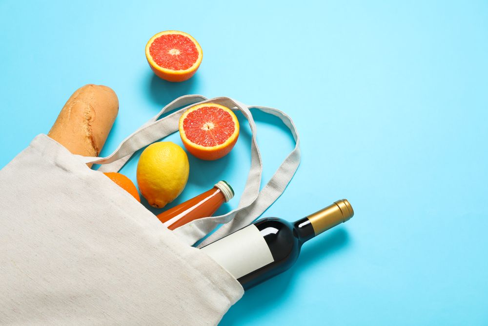 Eco bag with bottle of wine, juice, fruits and baguette on color background, space for text