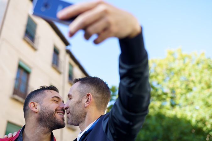 Two smiling men kissing and taking selfie outside