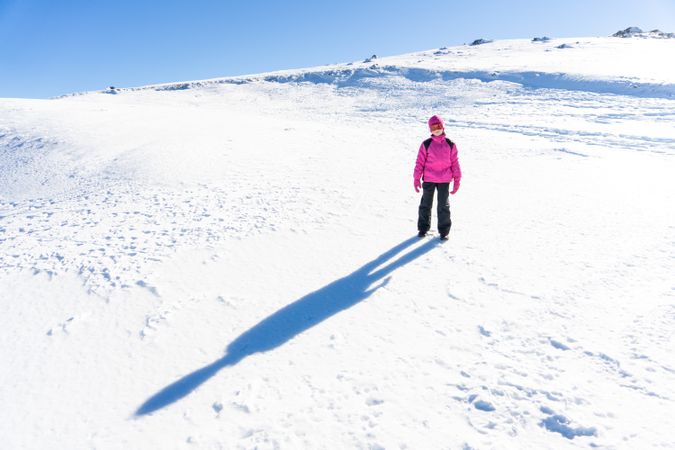 Child in pink snow suit standing with long shadow on snowy hill