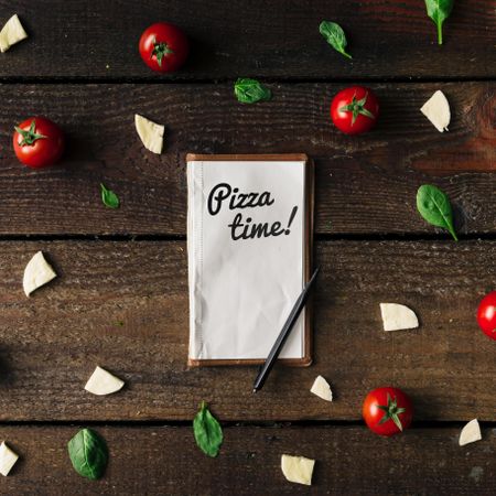 Basil, tomatoes, and cheese on wooden background with notepad and “Pizza time!” text