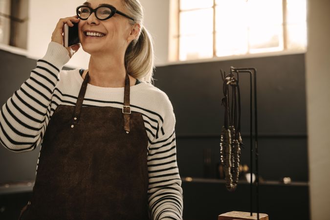 Portrait of happy woman wearing apron talking on mobile phone and smiling at jeweler workshop