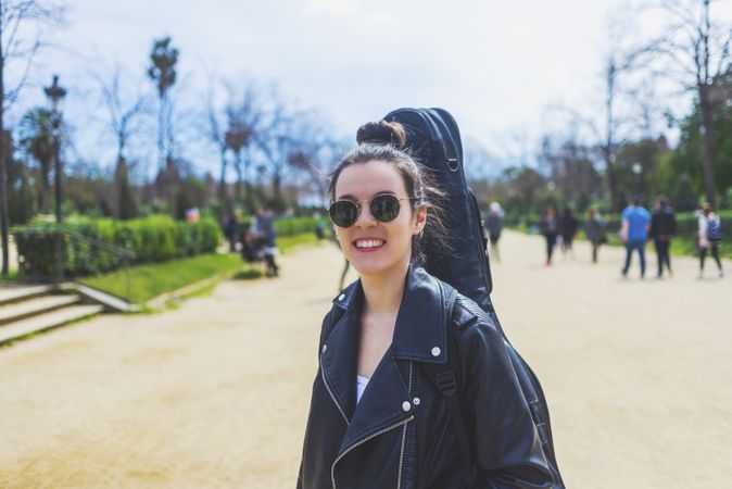 Front view of a young smiling hipster woman walking in a park in sunny day while carrying a guitar on back
