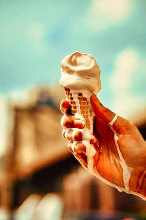 Hand holding melting ice cream cone against sky, vertical composition