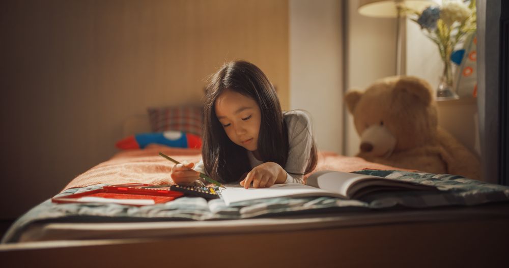 Portrait of a Little South Korean Girl Drawing with Colored Pencils in a Notebook While Lying in Bed in Children's Bedroom at Home.