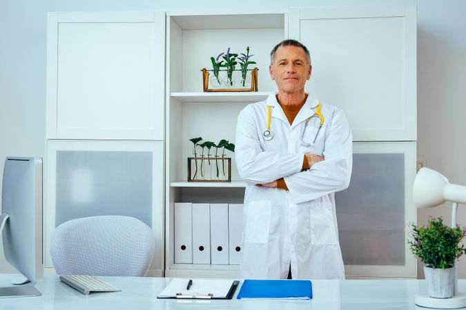 Mature male doctor standing in his office with arms crossed
