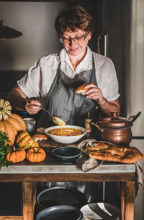 Woman with short hair and glasses with spoon and bread eating soup in cozy kitchen