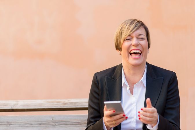 Laughing female in blazer sitting on bench holding phone in front of peach wall