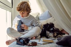 Attentive schoolboy with vintage photo camera near dog on bed