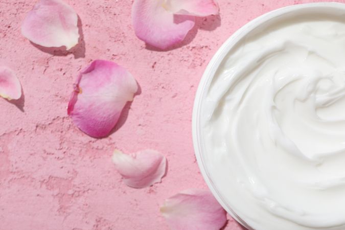 Jar with cream and rose petals on pink background, space for text