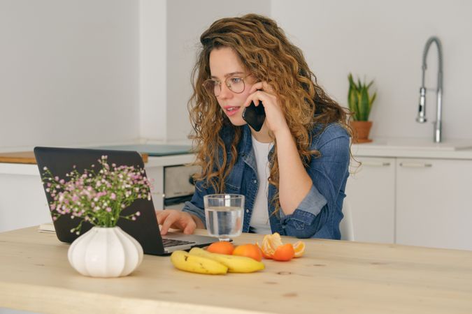 Young female freelancer talking on smartphone in kitchen
