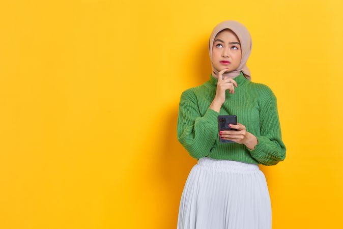 Woman in headscarf holding smart phone looking up while thinking about something