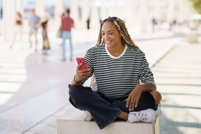 Happy woman checking mobile phone while sitting in pedestrian area outside