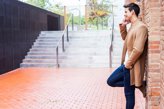 Side view of male in camel coat talking on phone while leaning on brick wall