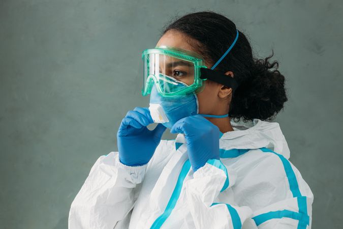 Side view of Black female medical professional in PPE gear adjusting her facemask
