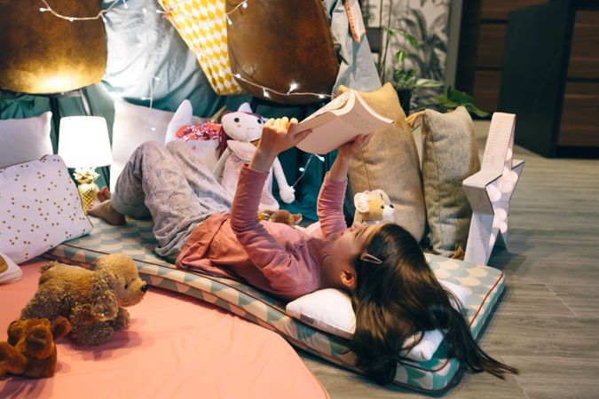 Happy little girl reading and relaxing lying over sleeping mat in tent surrounded by stuffed dolls