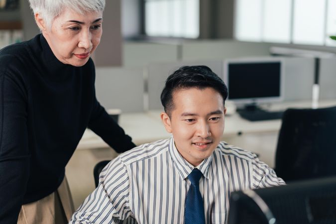 Serious mature Asian woman reviewing project on computer with male colleague