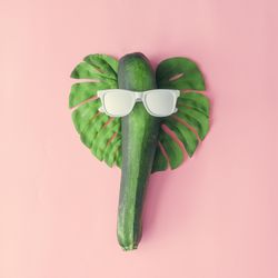 Monstera leaf, zucchini, and light painted sunglasses making face on pink background