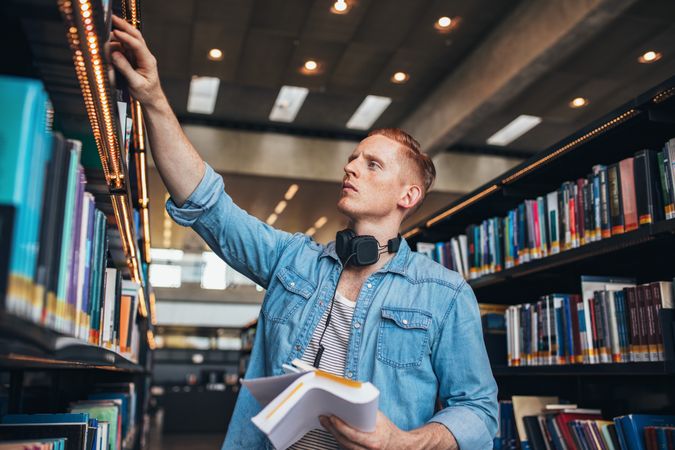 Shot of young male student selecting book from library shelf