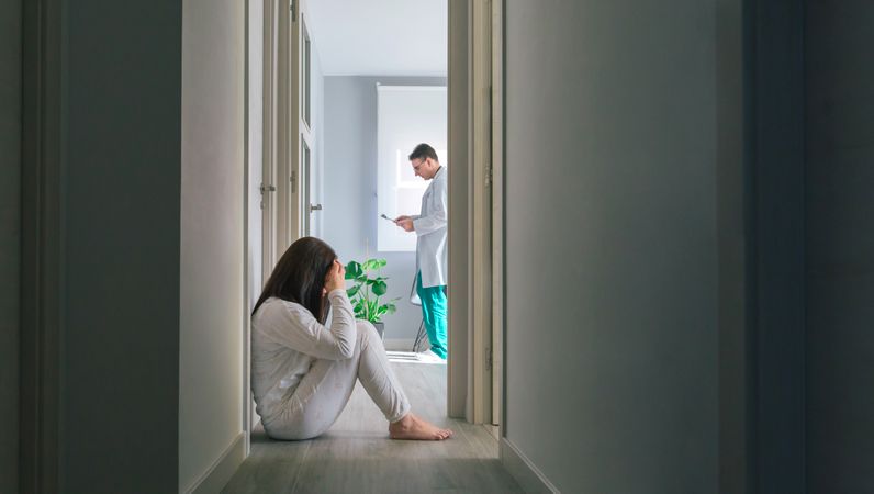Patient sitting on the floor in the corridor of a mental health center with a doctor in the background