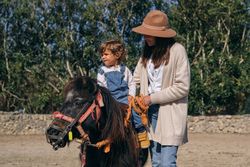 Smiling young woman standing near adorable toddler riding horse