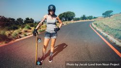 Full length portrait of young woman in helmet standing on a countryside highway with longboard 5qPKj0
