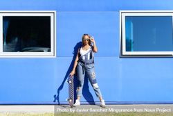 Female in denim overalls adjusting sunglasses in front of blue building 56Akj0