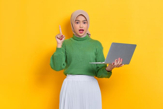 Surprised woman in headscarf holding up finger and holding laptop