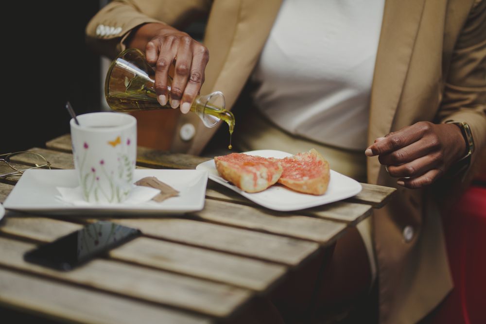 Black woman pouring oil on sandwiches at table with smartphone in cafe