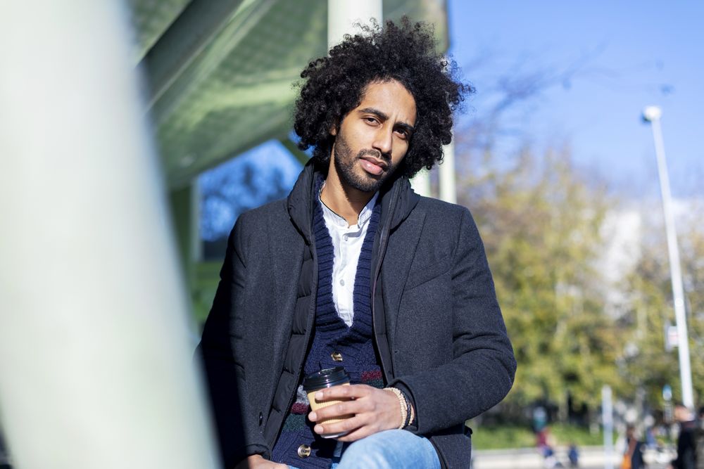Serious Black man in jacket with warm beverage balancing between poles outside