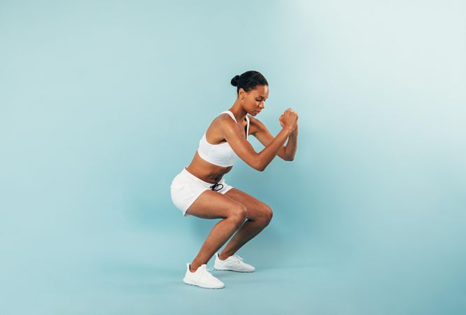 Fit woman in sports bra and shorts squatting in blue studio