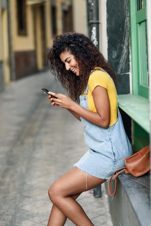 Smiling Arab woman perched on green window sill wearing denim dress while texting