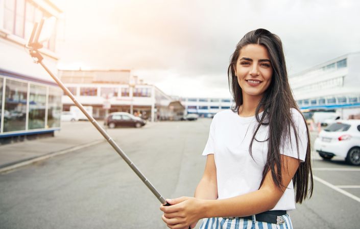 Woman with long brown hair standing in parking lot with selfie stick